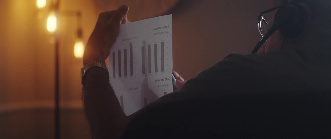 A man wearing headphones is using a pen to point to a paper with a graph while he is talking