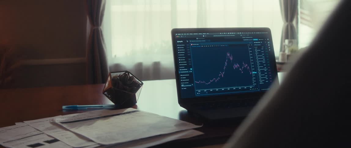 Close up of a laptop displaying a trading interface placed in front of a person seated at a table in a room