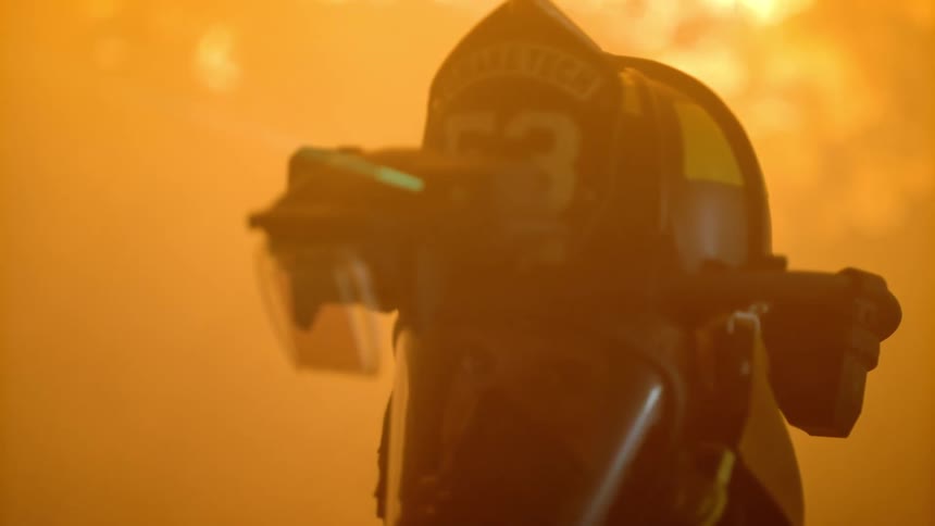 A firefighter wearing full gear stands outdoors at night with flames lighting up the surroundings