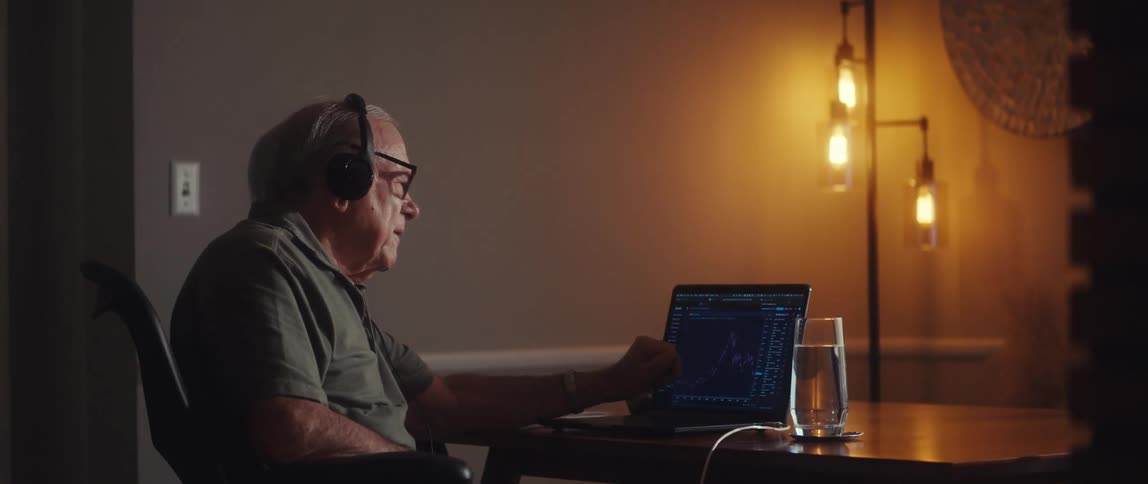 An elderly man wearing headphones analyzes stock charts on call at his desk