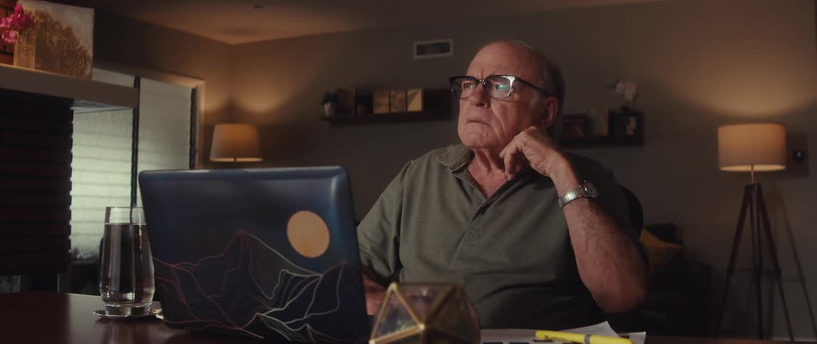Elderly man sits contemplatively at a table in front of a laptop in a living room
