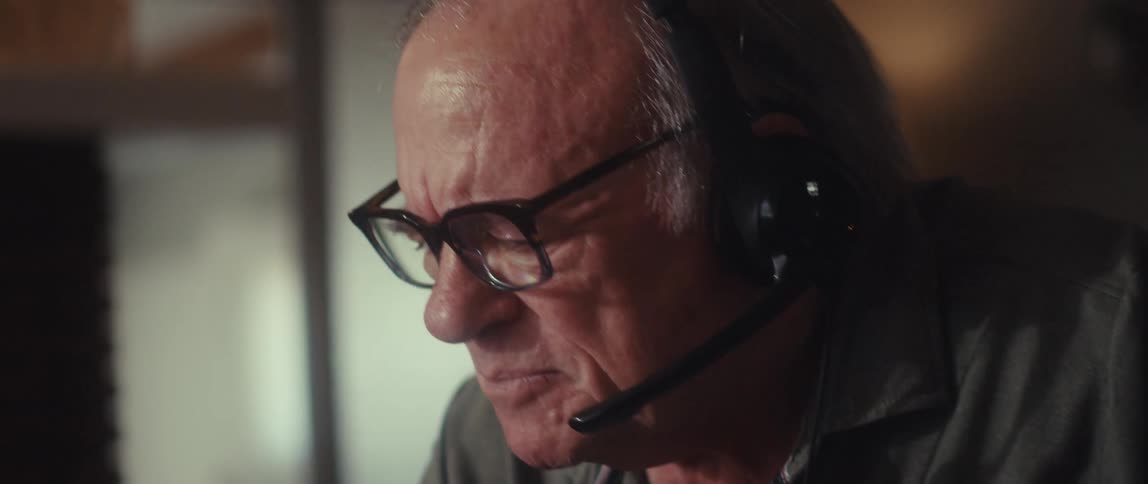 An elderly man is sitting in front of a computer wearing a headset