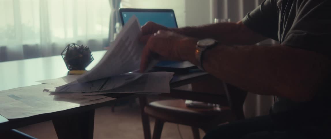 Elderly man sitting in front of a laptop using headphones and picking up documents