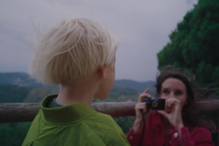 A woman and her son stand on a bridge high in the mountains as she takes a picture of her blondehaired child with a camera then gives him a kiss as the midday wind blows their hair