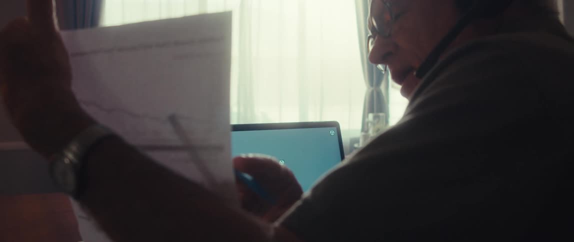 Elderly man with headphones presenting a document in front of a laptop