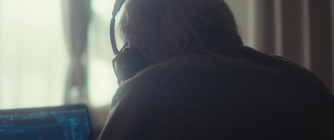 An elderly man sits behind a desk wearing a headset looking at a computer as he puts on his glasses to see the monitor clearly