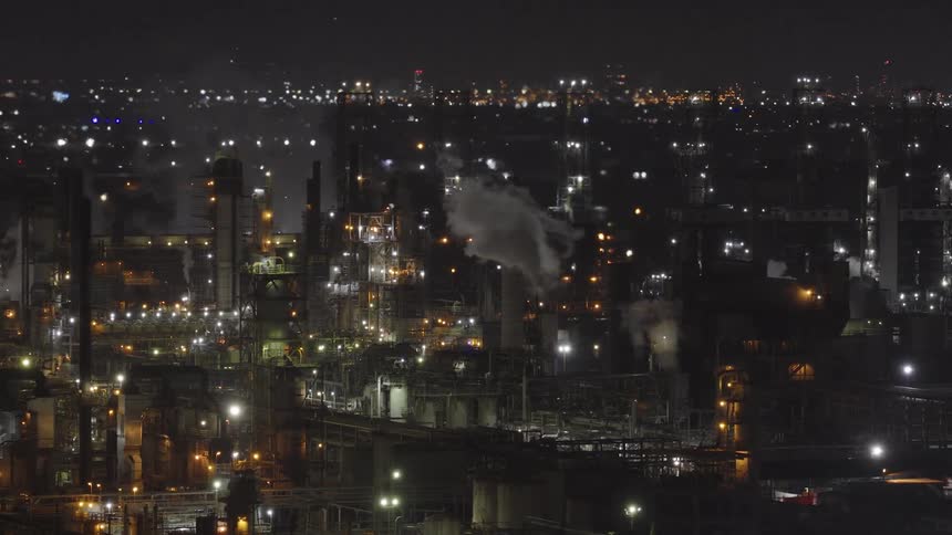 An illuminated industrial oil refinery billows smoke and steam into the night sky with a city skyline visible in the background