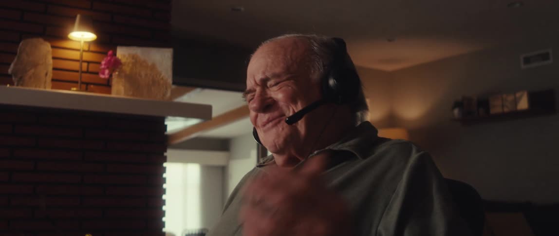 Elderly man holds a sheet of paper while speaking through a headset in a dimly lit room laughing as he gestures while explaining