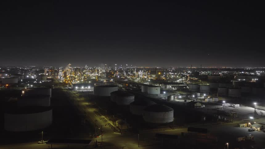 A large industrial refinery or oil storage facility glows with lights under the night sky