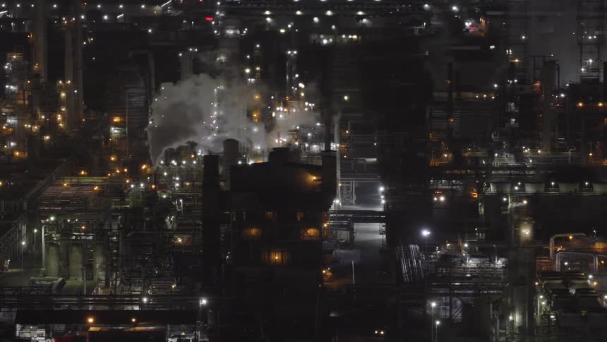 A slow aerial pan reveals a massive industrial refinery complex illuminated by thousands of lights at night as steam rises from its smokestacks