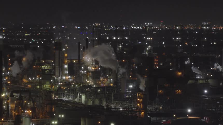 An illuminated industrial oil refinery emits smoke and steam into the night sky with a background of distant city lights