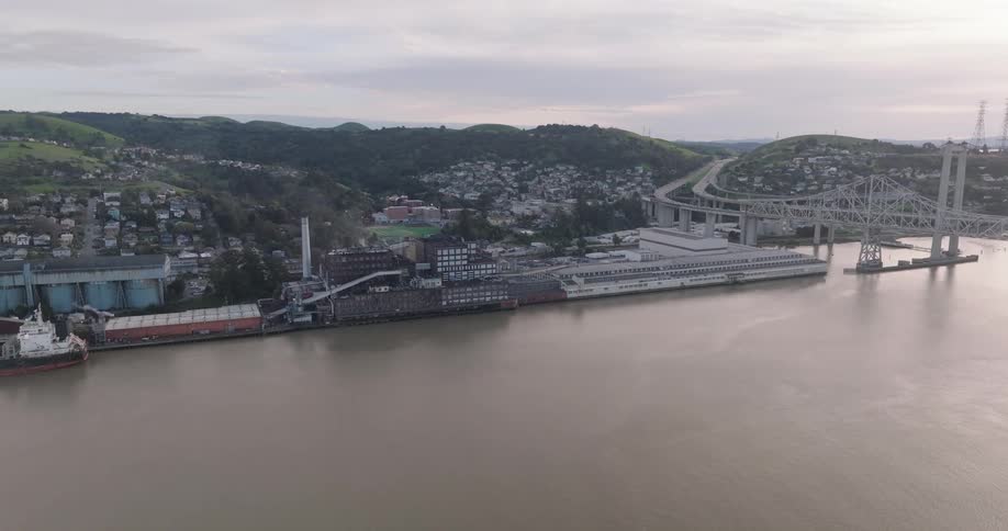 A cargo ship is docked at an industrial waterfront with a bridge and hillside homes in the background