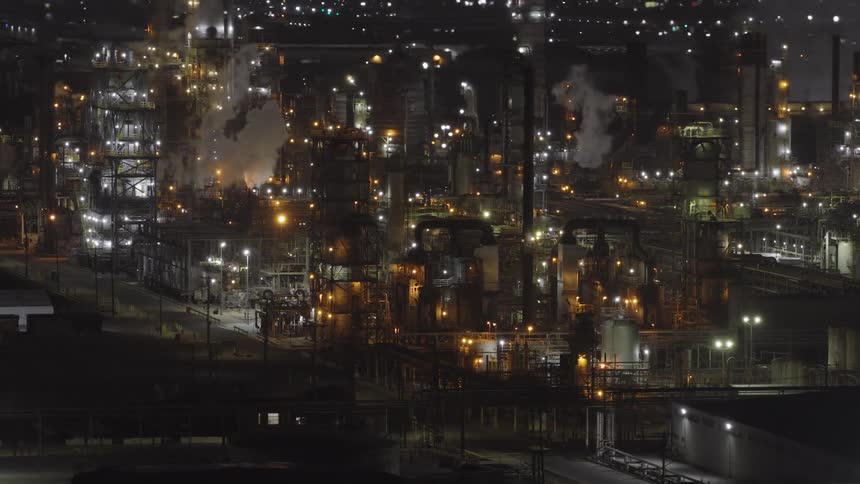 An illuminated oil refinery is captured from an aerial perspective at night with smoke and steam rising from its structures