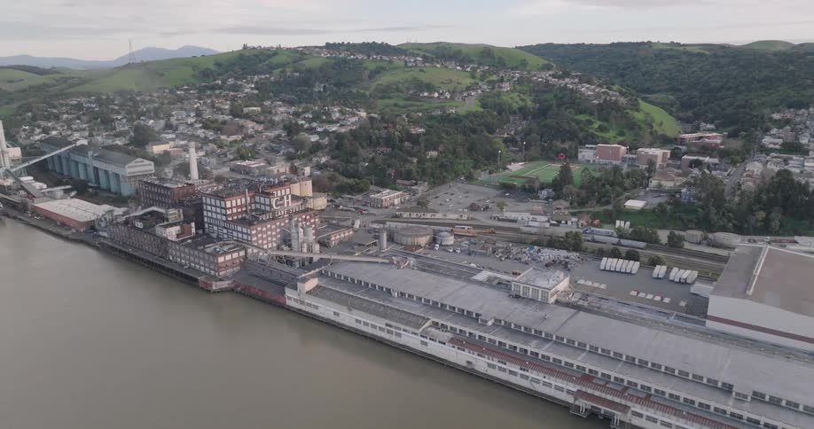 Aerial view of a waterfront factory with homes and green hills behind