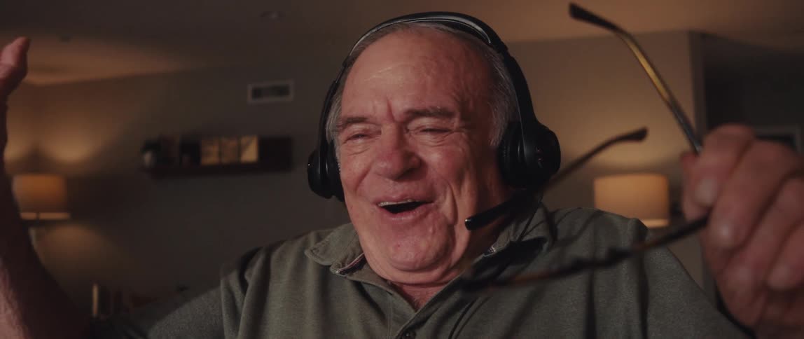 The face of an elderly man who is wearing headphones and he is sitting looking at a laptop and then he starts to get very happy and takes off his glasses while laughing