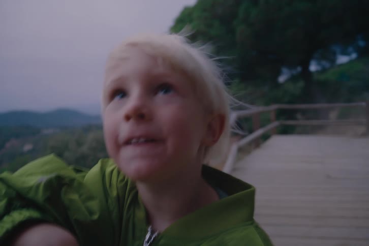 The face of a little blond boy wearing a green jacket and he is leaning on a wooden railing and looking to his side