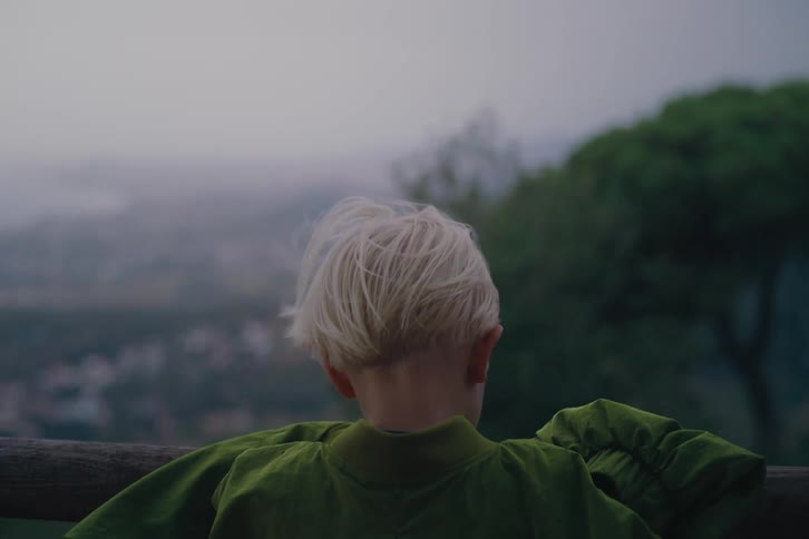 A kid wearing a green outfit stands on a bridge in the mountains at midday as the wind blows through his blonde hair