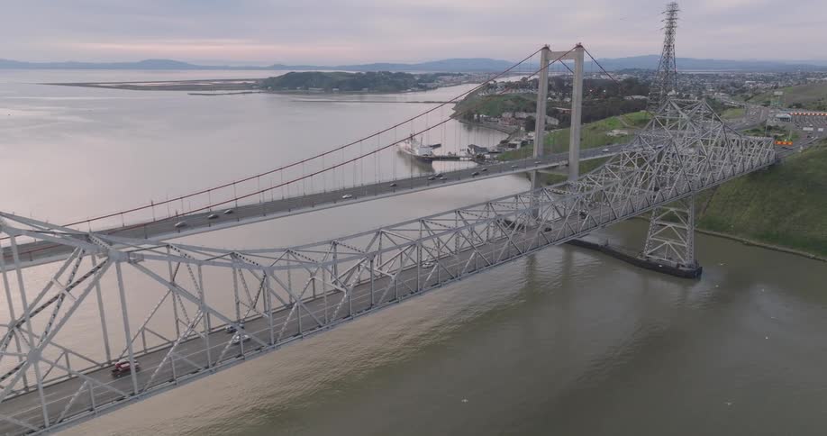 An aerial view shows a large metal bridge spanning across a body of water with land visible on both sides under a cloudy sky