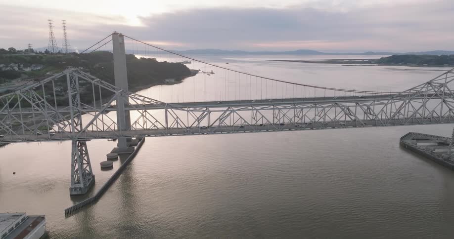 A large suspension bridge spans a wide river at sunset viewed from an aerial angle