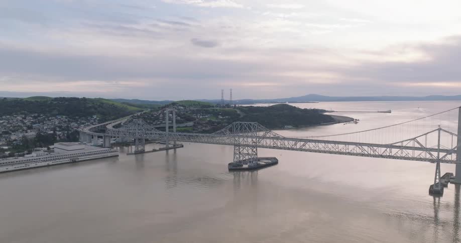 An aerial view captures the carquinez bridge spanning across the carquinez strait connecting land masses with urban development on one side and rolling hills on the other under a cloudy sky
