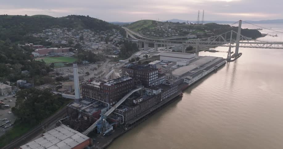 Cargo ships dock at a bustling industrial port with cranes warehouses and a bridge backed by hillside homes