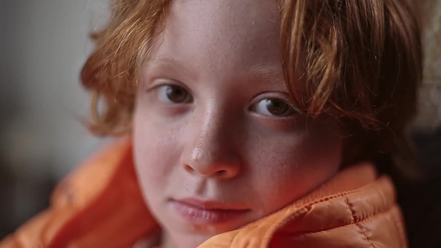 The face of a little blond boy staring straight ahead and he is wearing an orange outfit