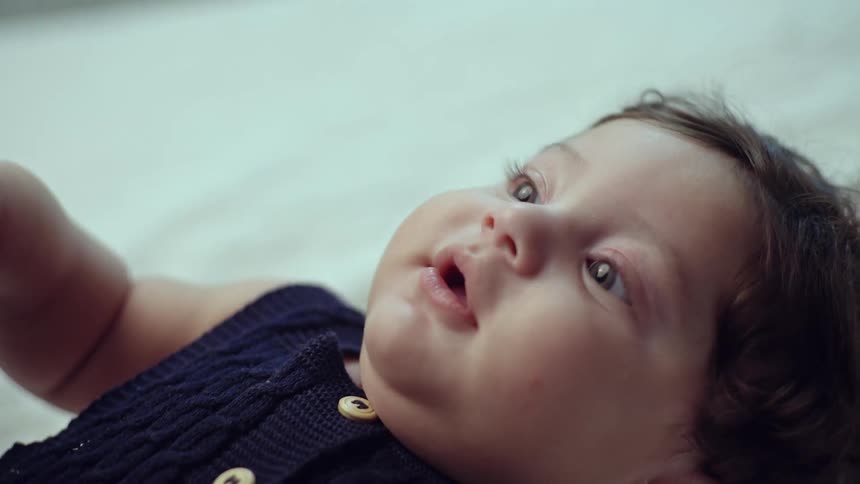 A close up shot captures a baby lying on its back looking up with wide eyes as a hand reaches out to touch its own