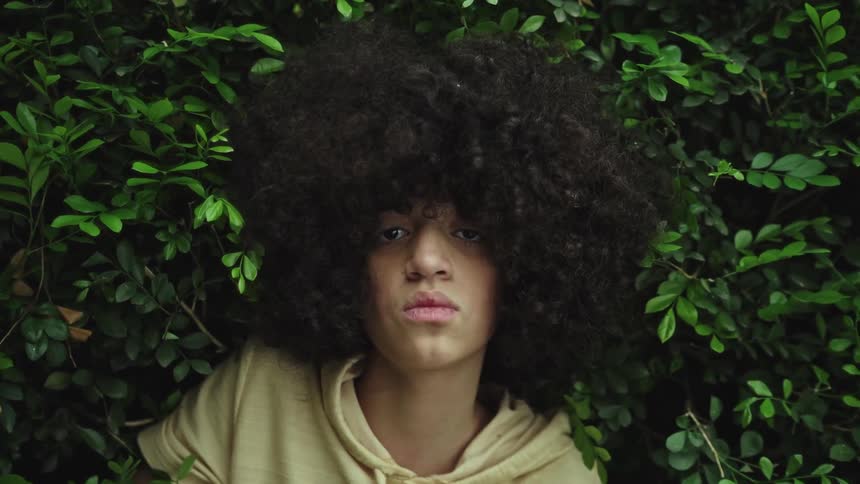 A teenager with an afro hairstyle and dark skin is surrounded by green leaves looking towards the viewer with a neutral expression