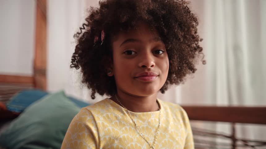 The face of a little girl wearing a yellow shirt is staring straight ahead and giving a subtle smile