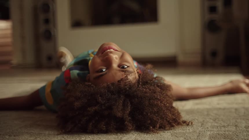 A smiling child lies upside down on the carpet indoors looking directly at the camera