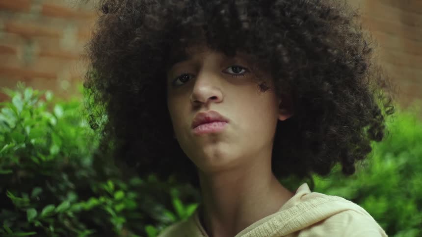 Curly haired youth stands near green bushes looking at the camera