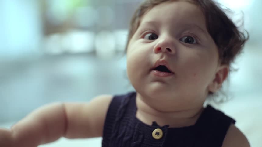 A baby with dark hair and light skin wears a dark blue top and looks forward with wide eyes and an open mouth