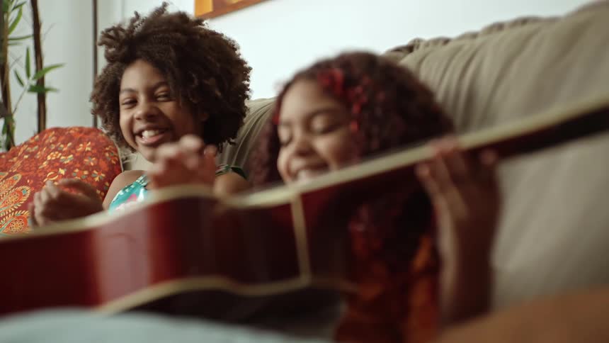 Two young girls relax on a couch as one explores playing a guitar