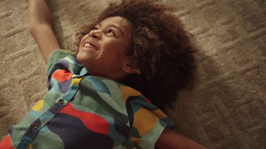 A smiling child lies happily on a carpet looking up at the camera