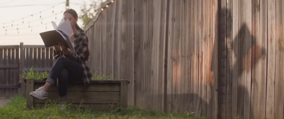 A young girl is sitting on a bench in a backyard and she is reading a book full of white pages