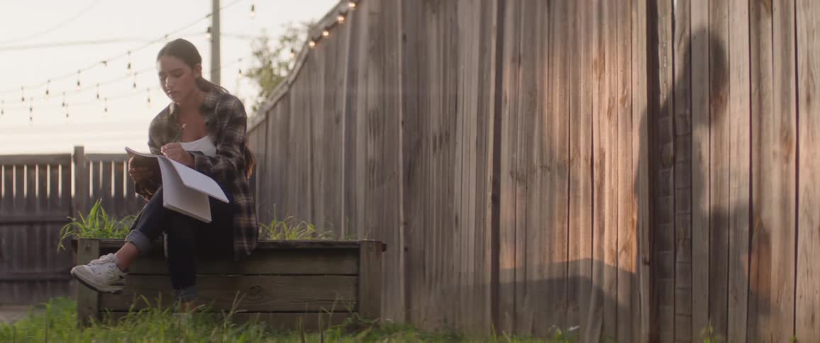 A young woman writes in a notebook outside by a wooden fence