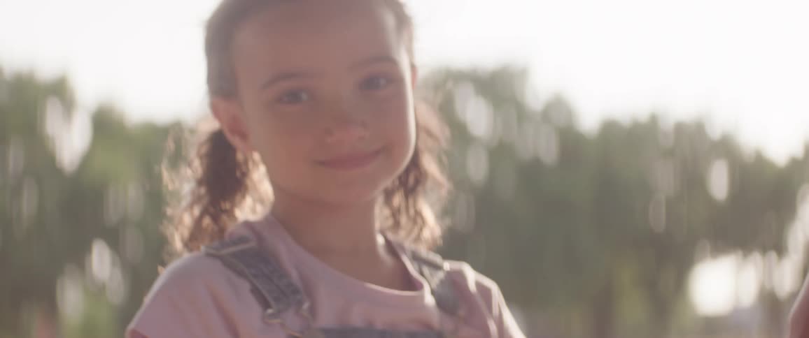 A portrait of a little girl who has a smile on her face and is looking straight ahead wearing overalls with a purple shirt