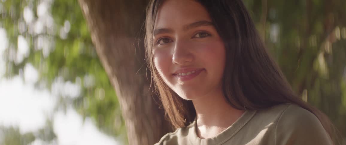 The face of a teenage girl who is smiling looking down in an outdoor environment and behind her there is a tree