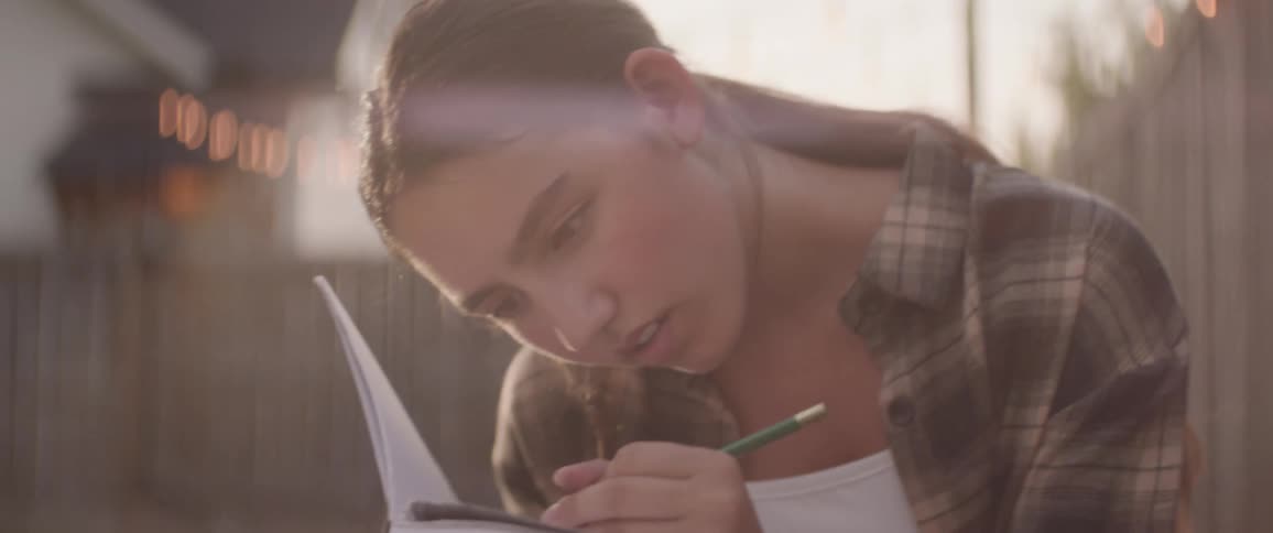 The face of a teenage girl who is writing on a paper using a pen and she is in an outdoor environment