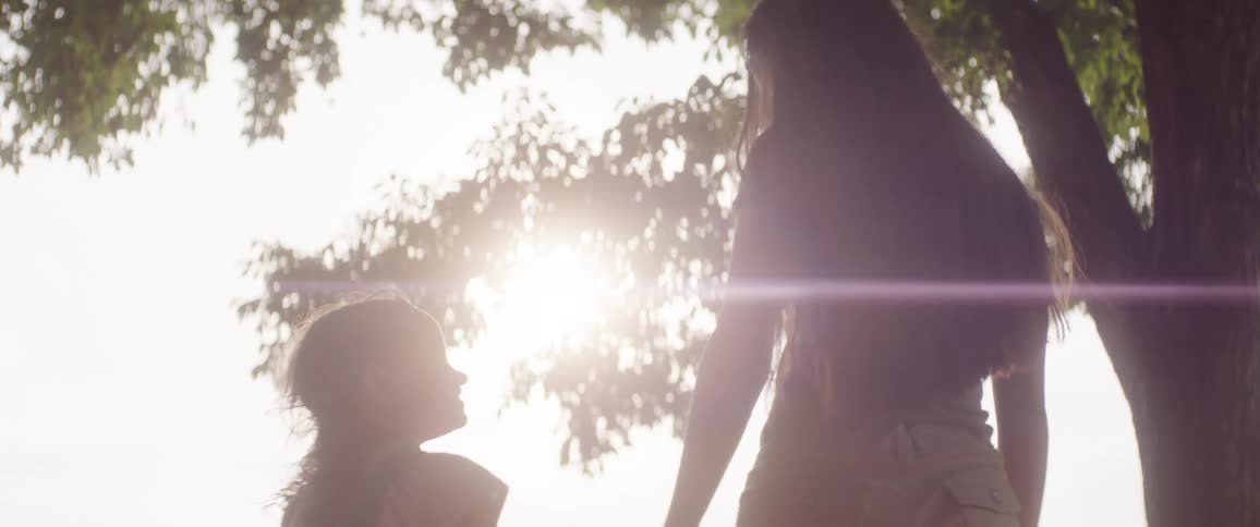 Female teenager happily holds hands with a girl with standing outdoors under a tree during daytime