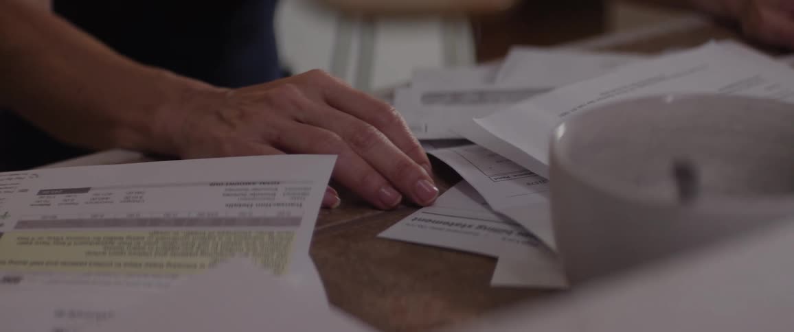 A brownhaired woman looks down anxiously at bills on the table her hands trembling as she struggles to stay composed
