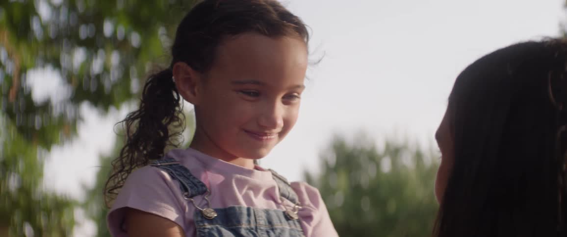 Two young sisters smile warmly at each other in a sunny outdoor setting