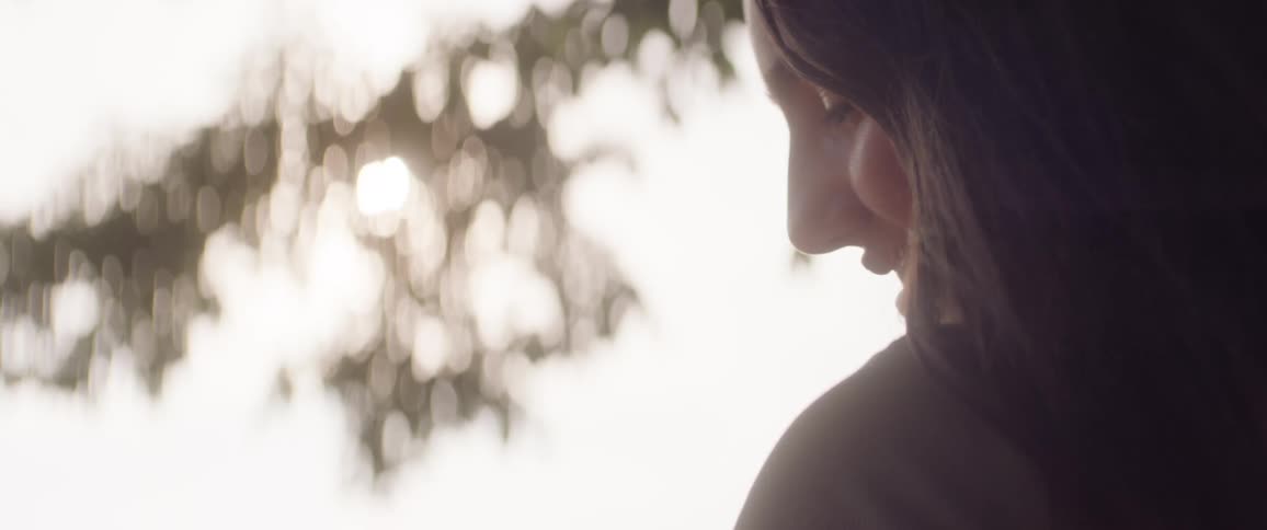 A young woman stands outdoors under a tree looking down with a smile on her face at midday