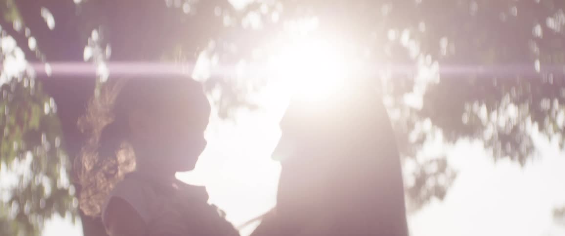 Mother holds her daughter while they smile at each other with sunshine filtering through leaves on them
