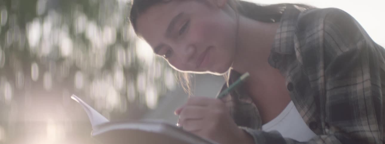 A teenage girl is outdoors using a green pencil to write on a paper