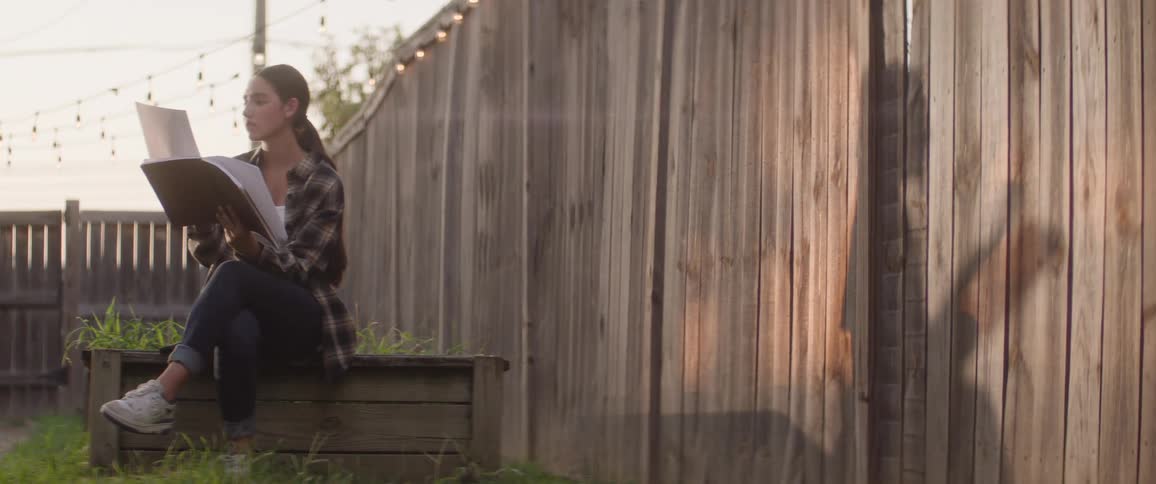 Female teenager goes through drawings in a sketchbook while sitting outdoors on a wooden structure beside a fence during daytime
