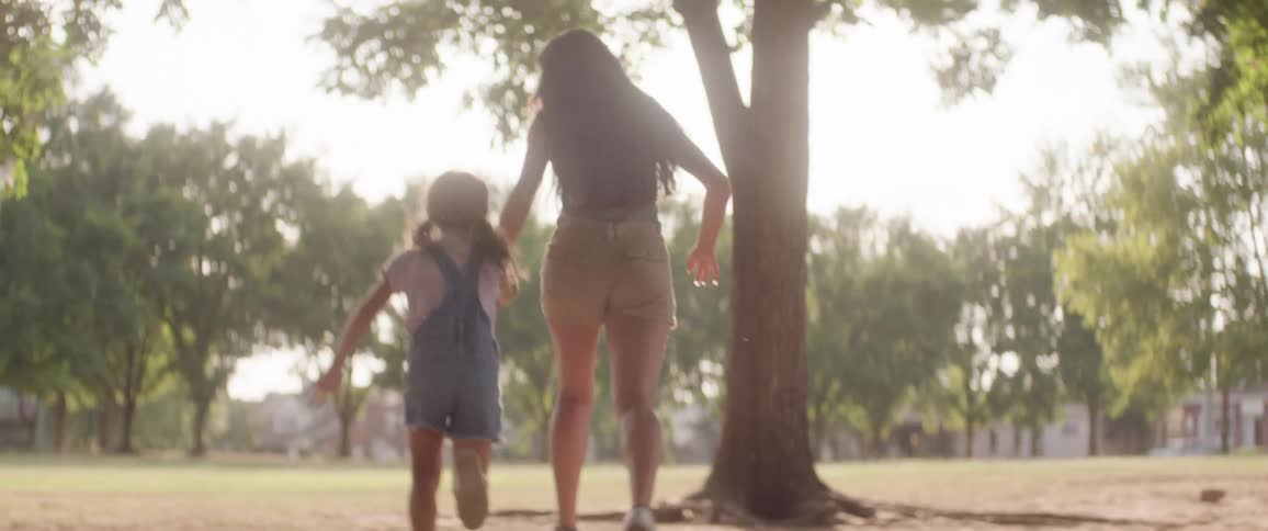 A woman and a girl hold hands in a field and run towards a small woodland area