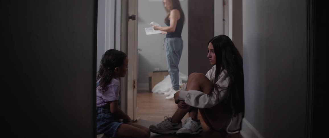 Two girls sit on the ground while a woman walks by