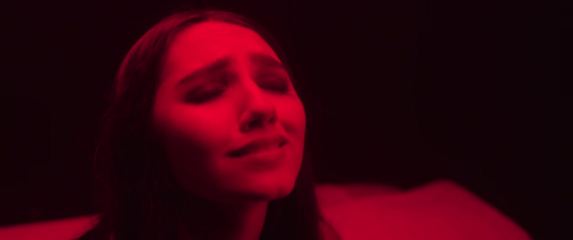 The face of a young woman who is looking up and then she closes her eyes and frowns and she is being illuminated by a red light