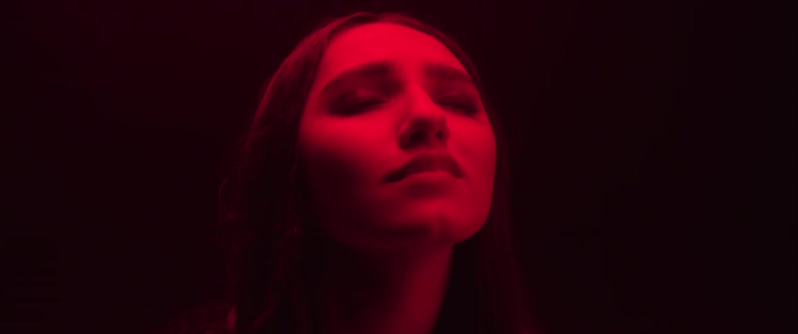 The face of a young woman who is looking up and then she closes her eyes and becomes sad and there is a strong red light illuminating her face